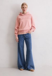 Różowa polarowa bluza z wytłoczonym logo, zestawiona z szerokimi niebieskimi dżinsami. Miękka faktura, luźny krój i warstwowe paski na mankietach. Brązowe obuwie.