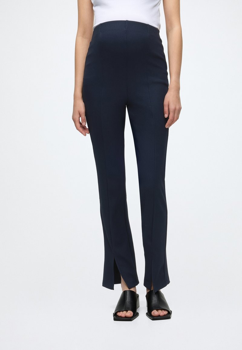 Personne portant un pantalon bleu marine taille haute avec des fentes à l'avant aux chevilles et des chaussures noires à bout ouvert à talon carré.