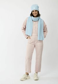 Roze katoenen tuinbroek met voorzakken, gecombineerd met een lichtblauwe gebreide sjaal en muts. Witte sneakers maken de outfit af.