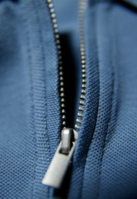 Zbliżenie na niebieską tkaninę z zamkiem błyskawicznym, wyposażonym w metalową pętlę do ściągania i zazębiające się ząbki, ukazującą gładką teksturę.