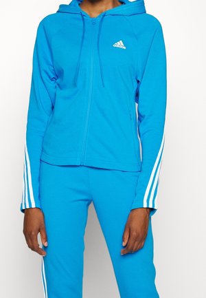 Personne portant un sweat à capuche zippé Adidas bleu vif et un pantalon assorti avec des bandes blanches sur les manches et les jambes.