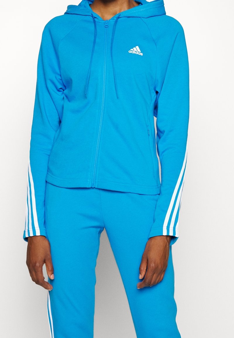 Personne portant un sweat à capuche zippé Adidas bleu vif et un pantalon assorti avec des bandes blanches sur les manches et les jambes.