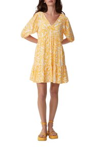 Gelbes Blumenkleid mit kurzen Puffärmelchen, tiefem V-Ausschnitt und gestuftem Rock. Kombiniert mit klobigen gelben Plateausandalen.