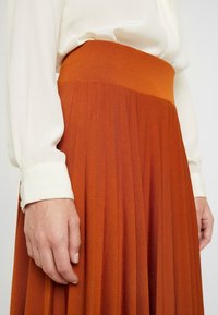Plisovaná oranžová sukně s vysokým pasem, vyrobená z texturované látky, v kombinaci s bílou blůzou s knoflíkovými manžetami.