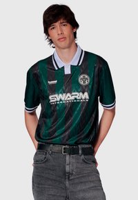 Grön- och svartrandig kortärmad fotbollströja med vit krage och logo med geometriskt mönster. Svarta jeans och läderbälte syns.