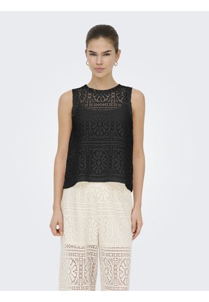 Kobieta ubrana w czarny, koronkowy top bez rękawów oraz białe, koronkowe spodnie z szerokimi nogawkami, stojąca na jednolitym tle.