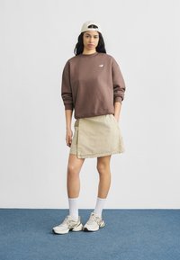 Καφέ φούτερ με ριμπ μανσέτες, μπεζ φούστα με πλαϊνή τσέπη και λευκές κάλτσες συνδυασμένες με γκρι και εκρού αθλητικά παπούτσια. Λευκό καπέλο φορεμένο ανάποδα.