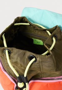 Sac à cordon vert et orange avec une fermeture éclair rose, doté d'un tissu doux et texturé, et une poche intérieure avec une étiquette en tissu.