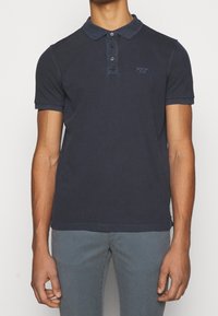 Polo-shirt bleu foncé avec une patte de boutonnage à trois boutons, des manches courtes et un petit logo brodé sur la poitrine gauche. Tissu en coton lisse.