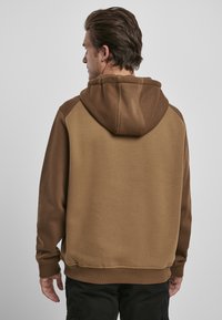 Brun hettegenser med en kontrasterende, lysere brun hette og skulderdetaljer. Glatt tekstur med en avslappet passform og ribbestrikkede mansjetter.