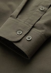 Olijfgroene katoenen shirtmanchet met twee grijze knopen, gladde textuur en nauwkeurige stikdetails. Heeft een afgeronde rand.