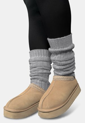 Beige Wildlederschuhe mit einer strukturierten beigen Sohle, kombiniert mit hellgrauen gestrickten Stulpen, die über schwarzen Leggings getragen werden.