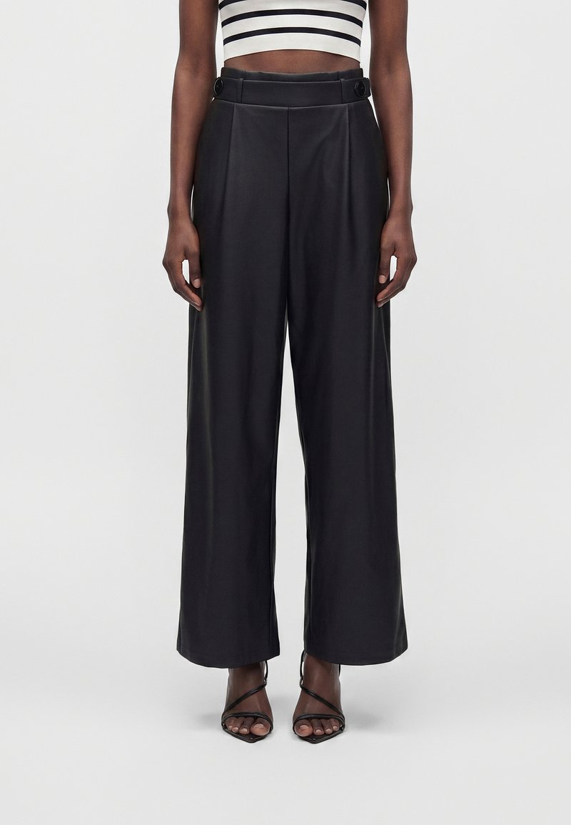 Pantalon noir taille mi-haute à coupe large avec détails de boutons à la taille, porté avec des sandales à talons noires à brides et un crop top rayé noir et blanc.