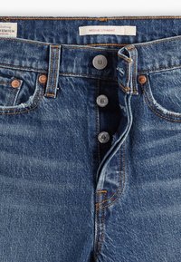 Jean en denim bleu taille haute avec fermeture à boutons, coutures orange, rivets métalliques et étiquette « WEDGIE STRAIGHT » à l’intérieur de la ceinture.