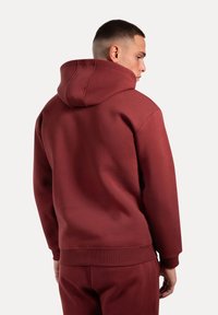 Felpa bordeaux con grande cappuccio con cordino, polsini e orlo a coste. Tessuto morbido, vestibilità comoda, con dettagli di cucitura visibili sul retro.