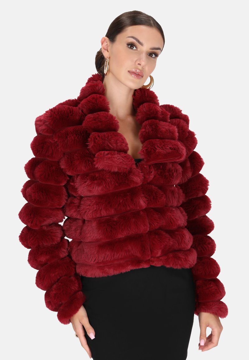 Frau trägt eine plüschige, tiefrote, strukturierte Pelzjacke mit großen Lagen, kombiniert mit goldenen Creolen und einem schwarzen Rock.