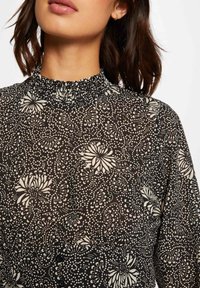 Schwarze Bluse mit einem hohen Rüschenausschnitt, floralem Muster in Weiß, aus strukturiertem Stoff mit zarter Transparenz.
