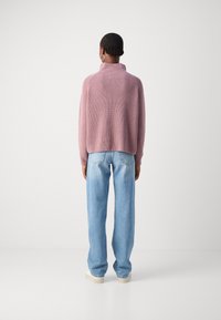 Pink knitted sweater with a high collar, ribbed texture, and cropped back, paired with relaxed blue jeans and white sneakers.