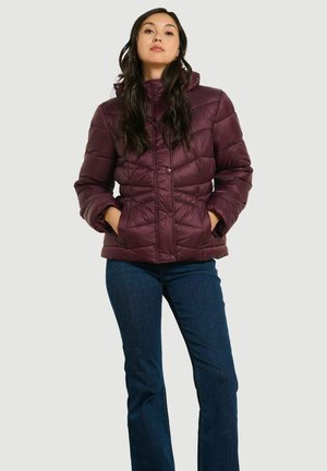 Donna con lunghi capelli scuri che indossa una giacca trapuntata di colore viola scuro e jeans blu scuro, in piedi con le mani nelle tasche della giacca.