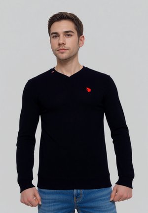 Zwart gebreid sweater met een V-hals, voorzien van een klein rood logo op de borst en een rode accent bij de kraag. Draagt met blauwe jeans.