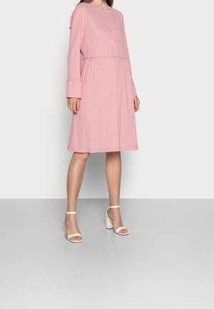 Femme portant une robe rose texturée à manches longues, longueur aux genoux, avec des manches retroussées et des sandales blanches à talons blocs, debout sur un sol gris.