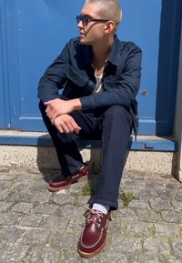 Bateaux en cuir marron avec des lacets blancs, portés avec un jean foncé et des chaussettes blanches, sur un fond bleu et une surface en pavés.