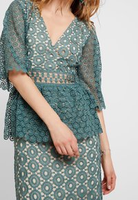 Teal spetsblus med djup V-ringning och utsvängda ärmar, med cirkulära mönster och peplumdesign. Matchande kjol med liknande textur.