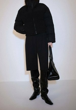 Persoon draagt een zwart sherpa-jas met rits, zwarte broek met wijde pijpen, zwarte slouchy kniehoge laarzen en houdt een grote zwarte handtas vast.