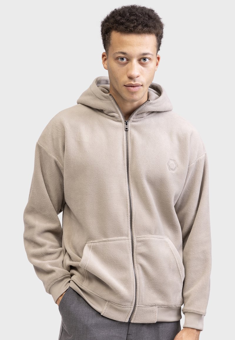 Sudadera de forro polar beige con cierre de cremallera, grandes bolsillos frontales y un logo hexagonal en el lado izquierdo del pecho. Textura suave y ajuste relajado.