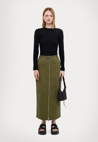 Maglietta nera a coste a maniche lunghe, gonna verde di media lunghezza con zip frontale e tasche, sandali neri e una piccola borsa nera.