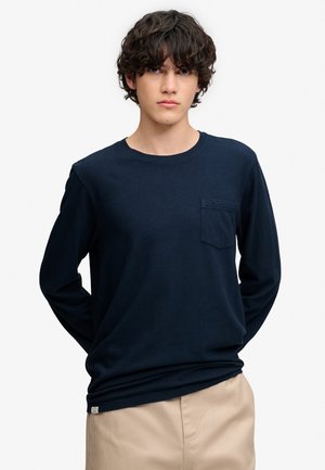 Ung mand med mørkt krøllet hår iført en navyblå skjorte med lange ærmer og brystlomme samt beige bukser mod en ensfarvet hvid baggrund.