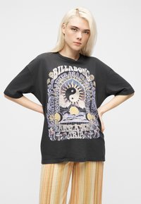 Νεαρή γυναίκα με ξανθά μαλλιά, φοράει μαύρο μπλουζάκι Billabong με σχέδια ήλιου, φεγγαριού, γιν-γιανγκ, και ριγέ κίτρινο-πορτοκαλί παντελόνι.