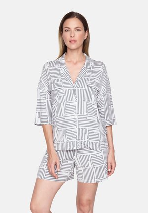 Vrouw gekleed in een wit-grijs geometrisch patroon overhemd met knopen en bijpassende korte broek, staand tegen een effen lichte achtergrond.