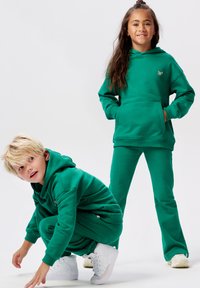 Twee kinderen in bijpassende groene hoodies en broeken; de een staat met de handen in de zakken, de ander hurkt, beide in witte sneakers.