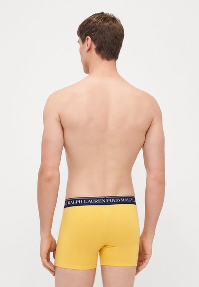 Rumene bokserice z mornarsko modrim pasom, na katerem je blagovna znamka "Polo Ralph Lauren", prikazane na osebi od zadaj.