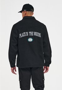 Chaqueta de lana negra con diseño de cuello, que presenta el texto blanco "PLACE IN THE WOODS" en la parte posterior y un parche circular verde. Mangas largas con botones.