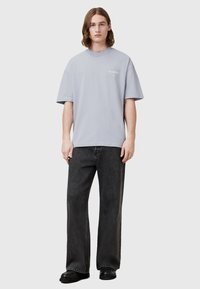 Tricou oversized gri cu guler rotund și detalii subtile cu logo, asortat cu blugi negri largi și cizme negre pe un fundal gri deschis.