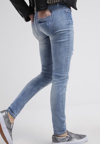 Světle modré džíny s úzkým střihem, s lehkými poškozeními a zadními kapsami, spárované se stříbrnými nazouvacími botami.