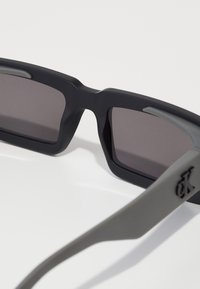 Ochelari de soare negri și gri cu un finisaj mat, formă rectangulară, lentile închise și detaliu cu logo pe braț.