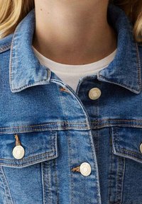 Blå denimjacka med klassisk krage, knäppt fram, två bröstfickor och synliga sömnadsdetaljer. En enklare vit halsringning under.