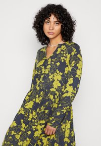 Vestido floral em azul marinho com padrão de flores amarelas brilhantes, mangas longas, botão na frente e design em camadas para uma textura adicional.