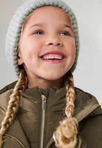 Fille avec deux tresses blondes portant une doudoune verte à col montant et un bonnet en tricot gris clair, souriant avec les dents visibles.