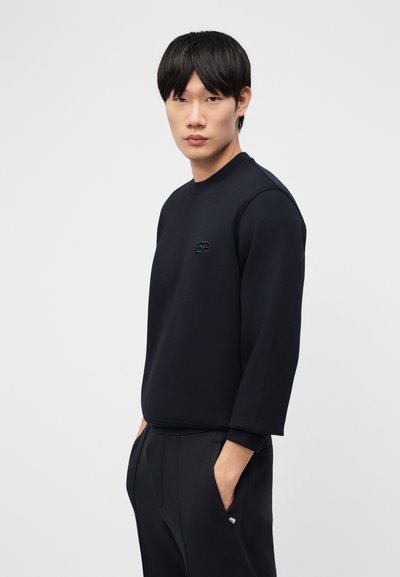 Νέος άνδρας με μαύρα μαλλιά, φορώντας μαύρο μακρυμάνικο φούτερ και μαύρο παντελόνι, στέκεται με τα χέρια στις τσέπες μπροστά σε απλό φόντο.