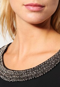 Haut noir avec un col orné de perles détaillées aux tons métalliques, présentant une texture lisse et une coupe ajustée.