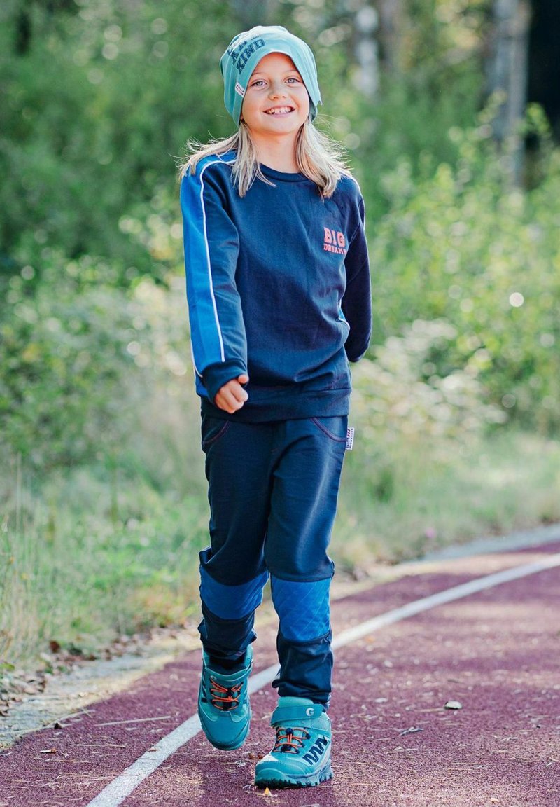 Blå sweatshirt med röd logotyp, matchande sweatpants och teal-beanie; blå och teal vandringskängor med orange snören; utomhus på spår.
