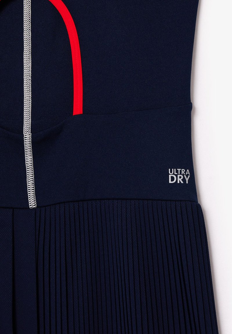 Mariene sportieve jurk met rode accenten, geplooide rok en "ULTRA DRY" label. Soepele textuur met contrasterende stiksel details.