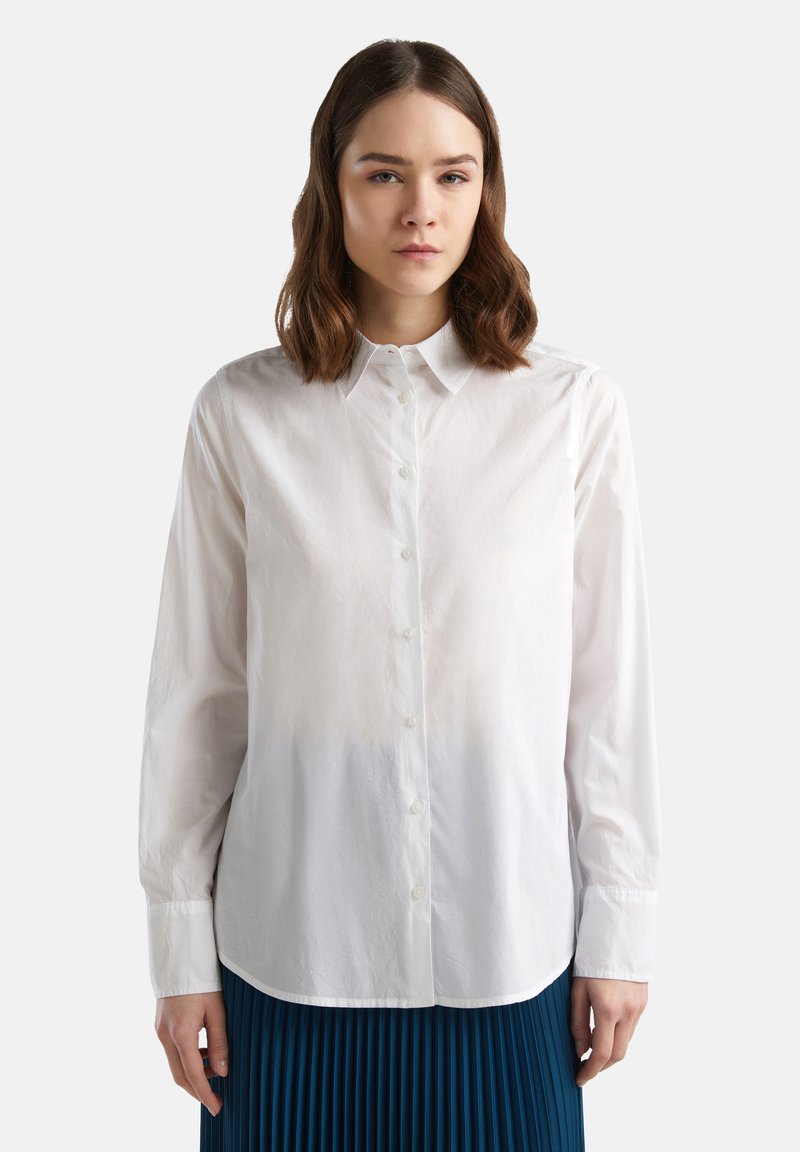 Camicia bianca a maniche lunghe con colletto button-down, texture liscia e orlo arrotondato. Abbinata a una gonna blu plissettata.