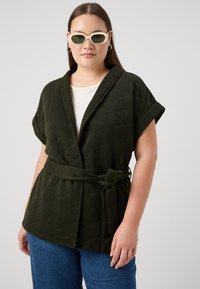 Tamsiai žalia siuvinėta apykaklė su diržu, trumpomis rankovėmis ir giliu V formos iškirpimu, derinama su šviesiai mėlynomis džinsinėmis kelnėmis ir žaliai nudažytais akinių saulės priekiniais stiklais.