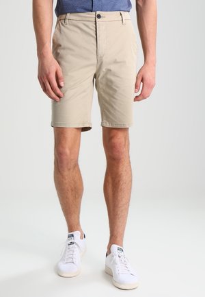 Mann trägt beige kniehohe Shorts, weiße Turnschuhe und ein blau gemustertes Hemd, steht vor einem einfarbigen hellen Hintergrund.