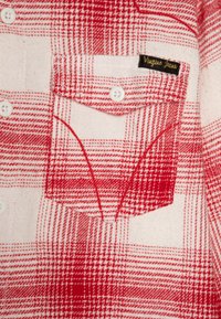 Rot-weißer kariertem Hemd mit einer Brusttasche, weißen Knöpfen und einem schwarzen Etikett mit der Aufschrift "Vigour Jeans". Weicher, strukturierten Stoff.